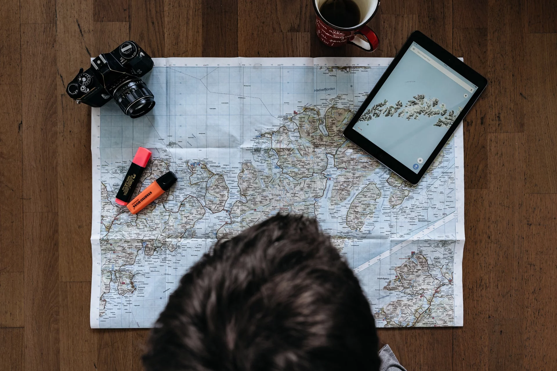 Fotografía cenital de una persona mirando un mapa sobre una mesa. Sobre el mapa hay una cámara fotográfica reflex, un par de marcadores y una tablet en la que también se ve un mapa.